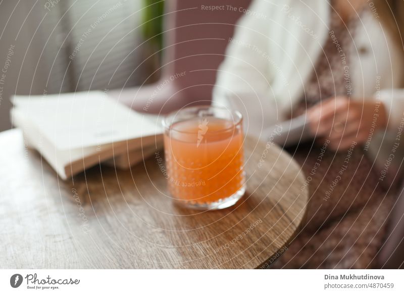 A cocktail of peach orange color on the table at cafe, restaurant or at home on wooden table. Lifestyle photo of woman reading ad drinking a refreshing juice drink.