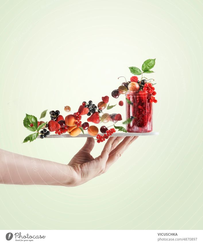 Women hand holding tray with red jam jar and various flying berries and green leaves women pale background creative food levitation concept above black currant