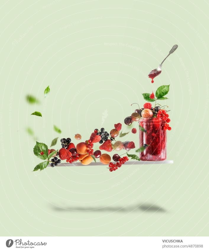 Flying cutting board with red jam jar, spoon and various berries at pale green background. Creative food flying currants strawberries raspberries cherries