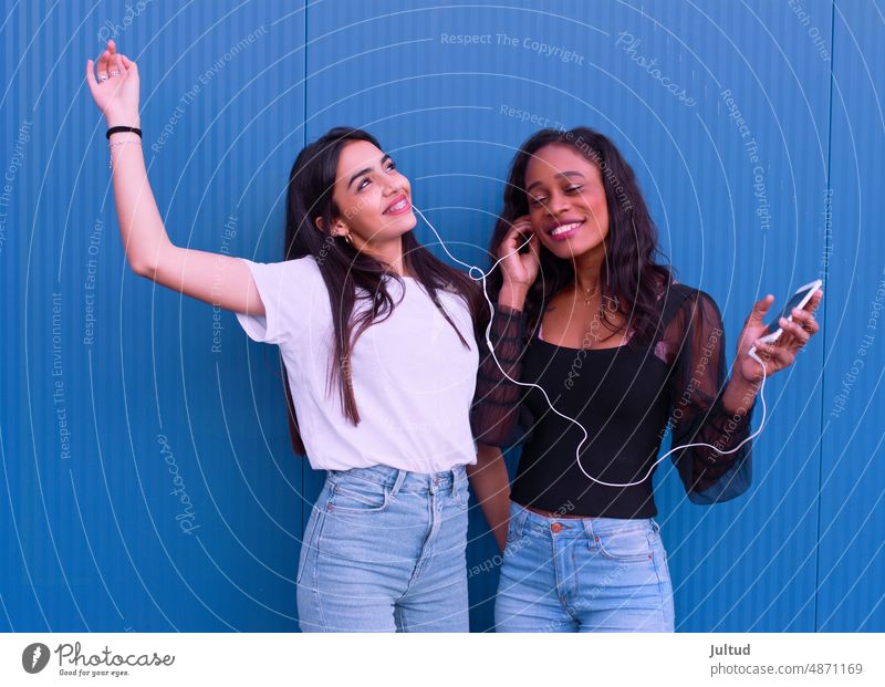 Two friends of different ethnicity listen to music in front of a blue wall. Girl Ethnicity spain Young Woman Twenties Trendy BuildingExterior Girlfriend