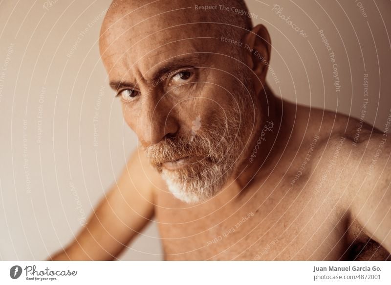 Bald man with bare torso and unkempt beard looks with serious expression, close-up and unfocused background bald naked disheveled left mustache red-haired