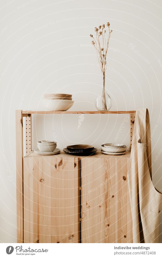 A wooden sideboard cabinet with ceramic plates and bowls, dried flowers in glass vase and a cotton apron on it. Minimal home decoration layout idea. Pastel and brown tones.