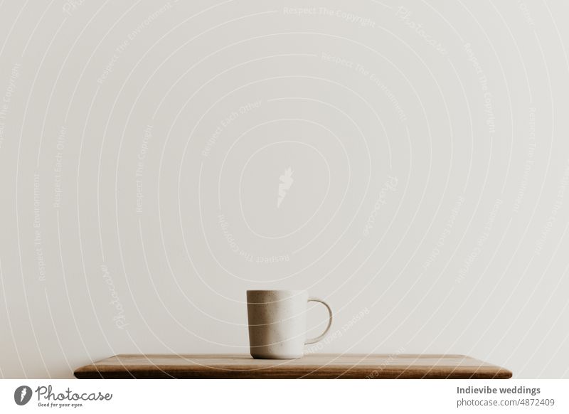A beige ceramic mug on a brown wooden board in front of a white wall. Hand made coffee cup with copy space. Minimal concept, simple design. tea cafe isolated