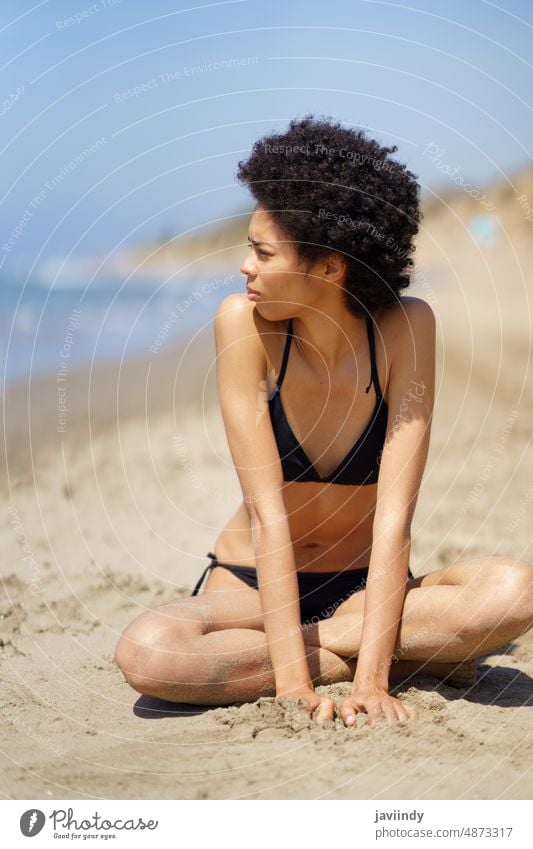Black woman in bikini sitting near sea beach shore summer swimwear seacoast chill sunny leisure recreation trip seashore female sand black ethnic legs crossed