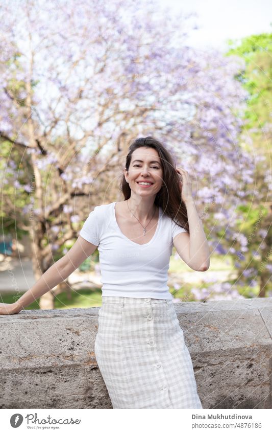 Young female brunette tourist posing near jacaranda tree, smiling. Neutral colors in clothes style. Beige color walls. Woman tourist wearing white t-shirt.