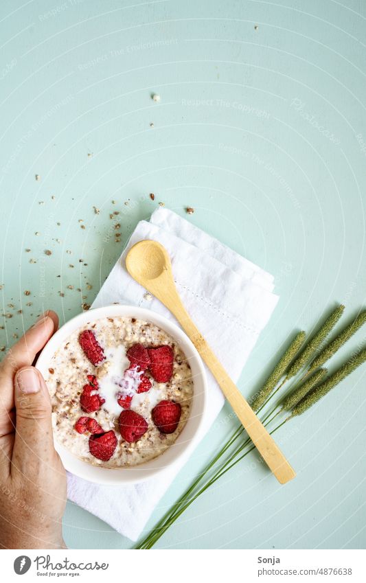 Hand holding warm porridge with fresh raspberries in a bowl on a table. Porridge. Breakfast Raspberry Milk Bowl Woman Oat flakes Spoon Table Plan Food Cereal