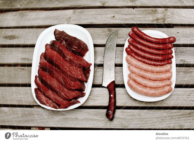 A plate of beef rump steaks and a plate of bratwurst and merguez sausages stand side by side on a wooden table, with a large wooden-handled meat knife in the center.