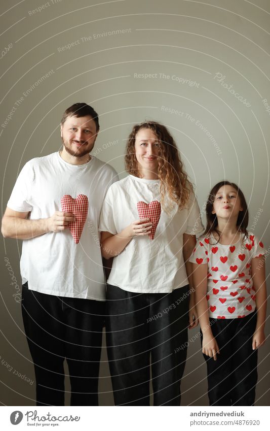 valentines day. happy family, mom, dad and little daughter in white t-shirts holding hand made red hearts in their hands looking at the camera on white background. happiness, health and love concept