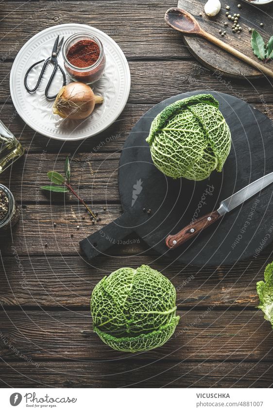 Whole raw savoy cabbage at wooden table whole black cutting board knife onion spices kitchen utensils cooking home seasonal healthy ingredients top view