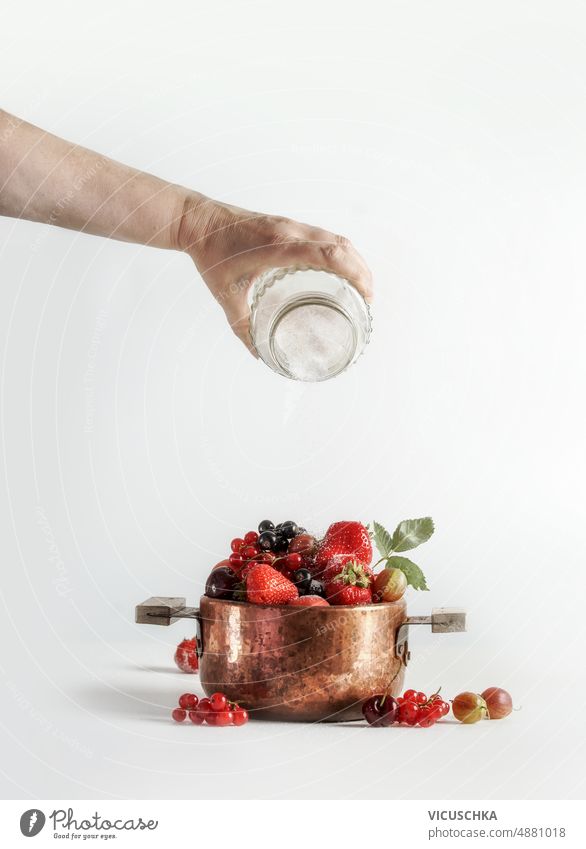 Jam preparation with woman hand hand pouring sugar on copper cooking pot with various summer berries and fruits at white background. jam above black currant