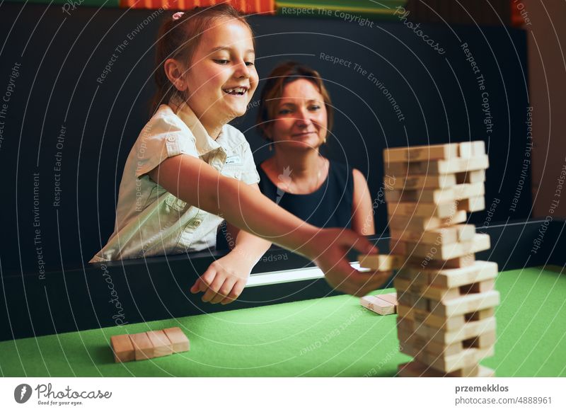 Excited girl playing jenga game with her mom in play room. Girl removing one block from stack and placing it on top of tower. Game of skill and fun. Family time