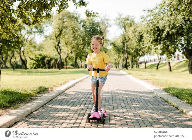 An active little girl rides a scooter on a path in an outdoor park on a summer day. Seasonal children's active sport. Healthy lifestyle in childhood outdoors