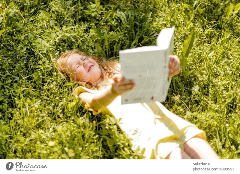 A charming baby is lying on the grass in the garden or park with a book and squinting from the sun girl school knowledge summer read a book summertime sunny