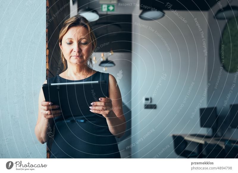 Businesswoman working on tablet in office. Mature woman using touch pad computer standing by window in modern interior. Manager focused on work holding digital device. Using technology