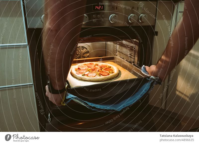 A man bakes pizza at home using an oven. Men's hands pull a pizza baking sheet out of the oven. male cook make pepperoni jalapeno hot pepper chili pepper pan