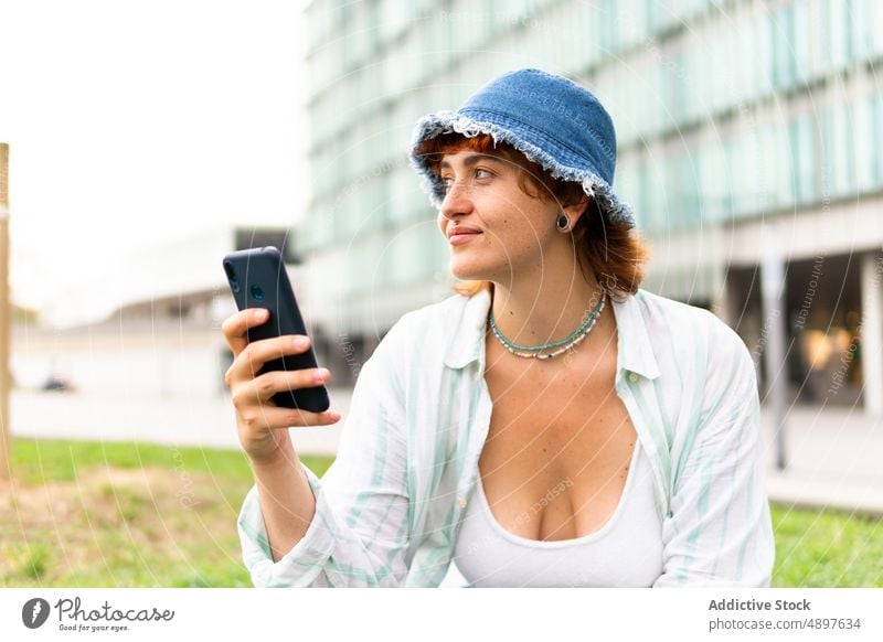 Smiling woman browsing smartphone on lawn online internet street city chat social media building female surfing grass cellphone positive glad style pastime