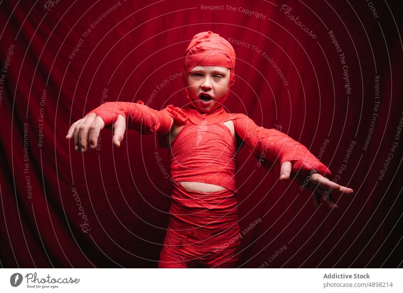 Cute little kid in mummy costume shouting and gesturing in red studio child halloween boy scary portrait spooky reach out make face creepy adorable celebrate