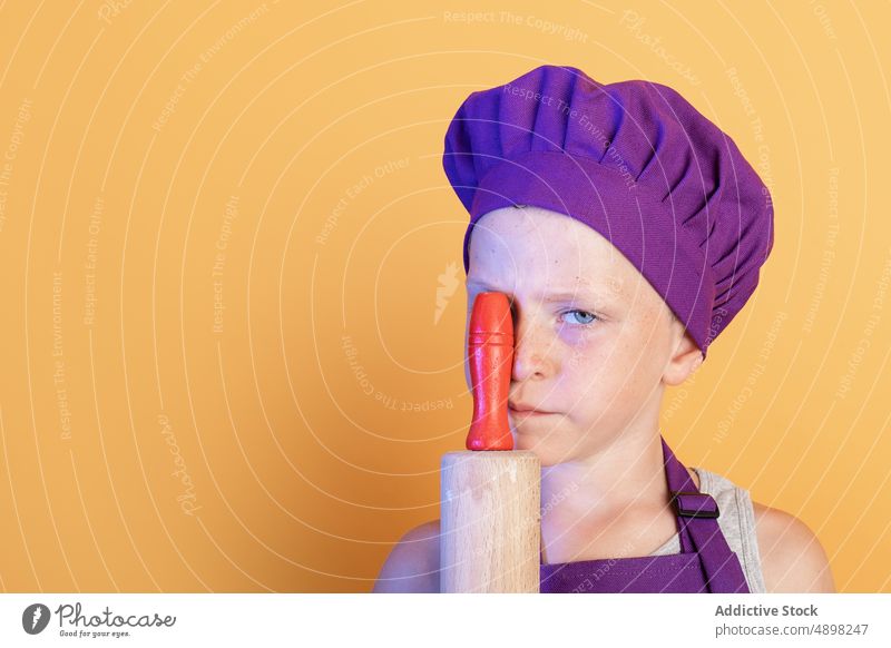 Cute little child holding rolling pin near face and looking at camera boy chef funny playful cover eye kitchenware portrait grimace cook childhood culinary