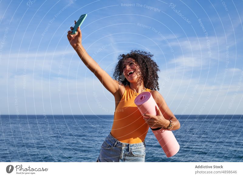 Cheerful Hispanic woman with mat taking selfie on seashore yoga healthy lifestyle training hobby self portrait social media smartphone cellphone mobile capture