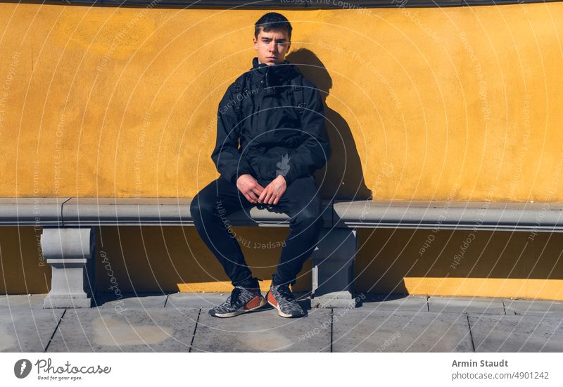 young man sits on a stone bench with yellow wall in the background sitting serious relaxed break backpack journey portrait teenager looking male beautiful