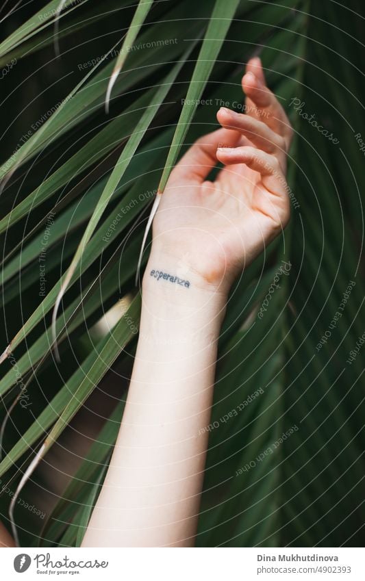Hand relaxed with green palm leaf as background with word "Hope" in Spanish. Tattoo art on a wrist of female's hand in summer. Touching tropical nature. Concept of nature and human living together. Eco-friendly concept. Tropical beauty.