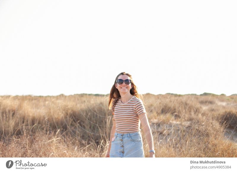 Happy young woman wearing sunglasses walking near the beach in dunes with tall dry grass. Photo in neutral beige colors. traveler beautiful landmark tourism