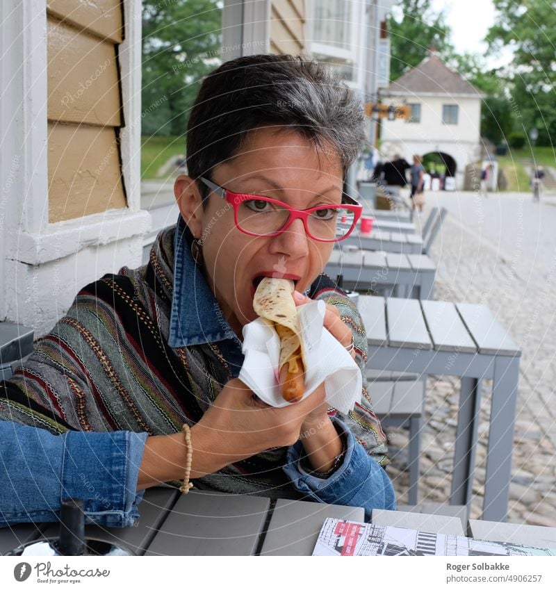 Woman eats sausage in a tortilla, at an outdoor restaurant Color Sausage sandwich Tortilla Street table Outdoor restaurant Eating Lady Red glasses Black hair