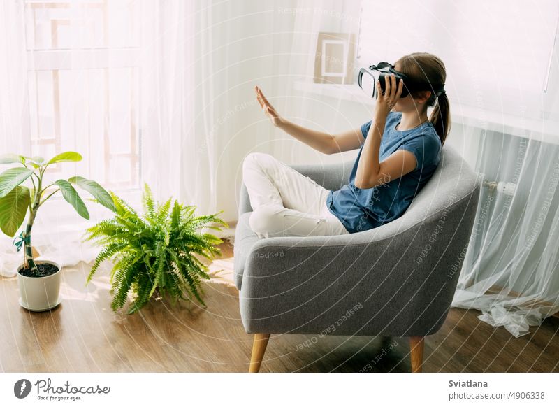The girl is sitting in a chair with virtual glasses studying online or playing virtual games. Modern technologies, virtual learning, modern leisure and education