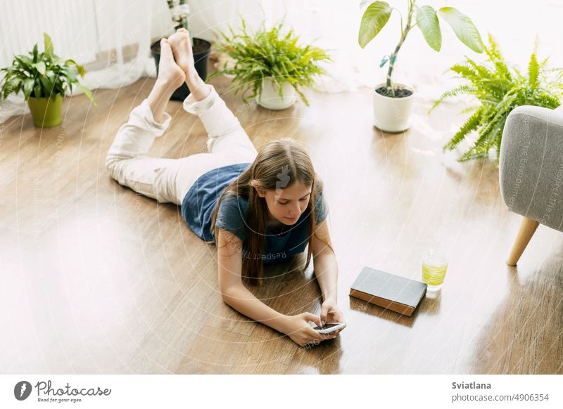 A happy teenager is texting on a mobile phone, lying on the floor in the living room. Modern technologies, online communication student girl technology message