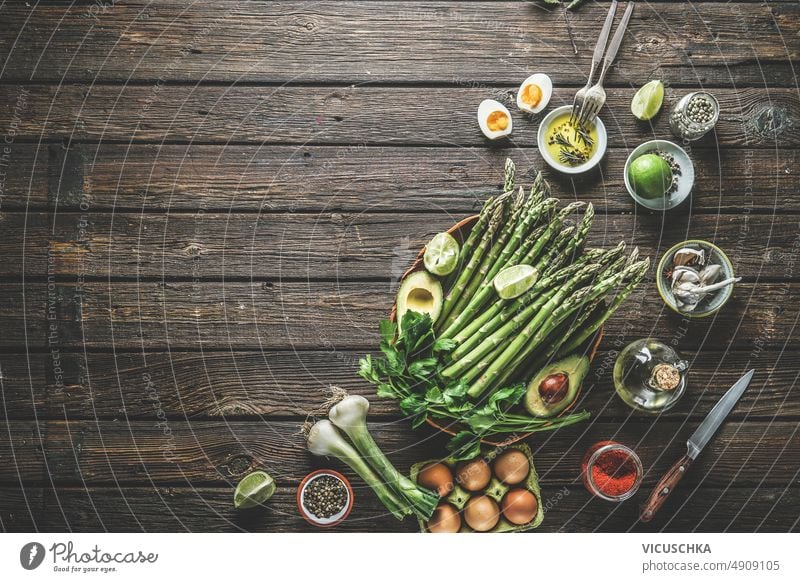 Food background with green asparagus and various healthy ingredients: avocado, egg, spring onion, lime, spices and herbs at dark rustic wooden kitchen table