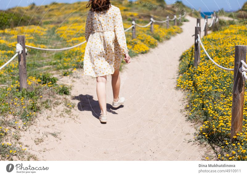 Young woman in sundress from behind on the beach among blooming dunes with yellow flowers. Photo in neutral beige colors. traveler beautiful landmark tourism