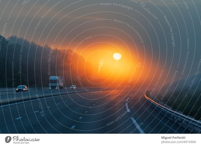 Landscape of a highway with a truck and several vehicles circulating at dawn, and the sun appearing through the clouds, allowing its circumference to be seen through the mist.