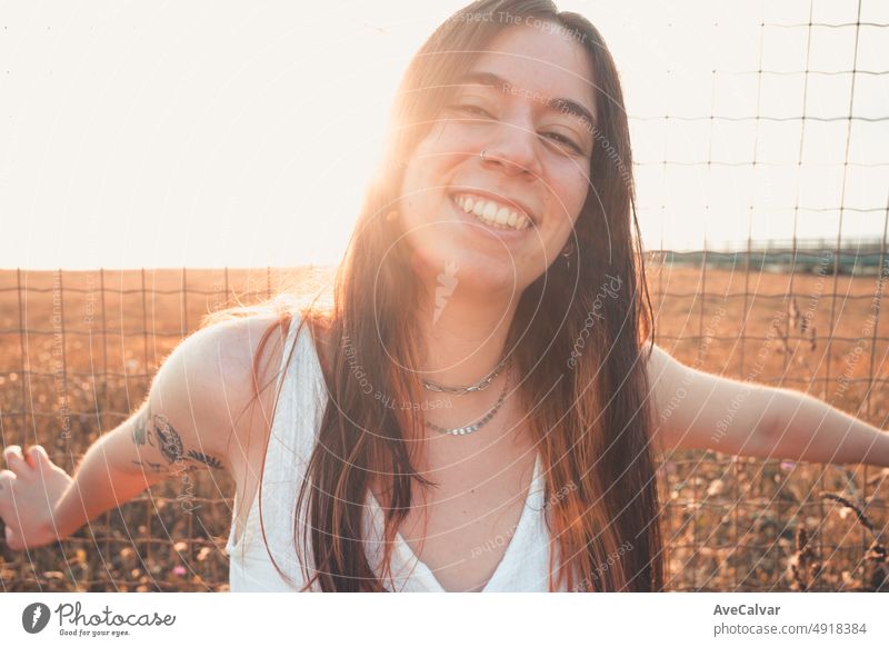 Super happy and positive smiling young woman with tattoos resting against a fence while closing eyes relaxing. Carefree holiday free time concept. Freedom and liberty concept. Gen Z outdoors fun.