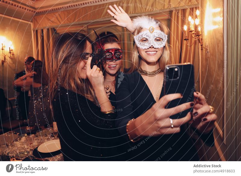 Positive women in carnival masks taking selfie in restaurant self portrait dinner masquerade friend having fun social media occasion bonding pastime enjoy