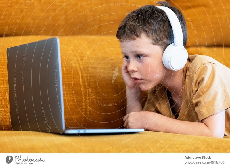 Boy sitting on a sofa with headphones on his head and using the laptop child computer boy caucasian technology male kid serious concentrate cartoon movie