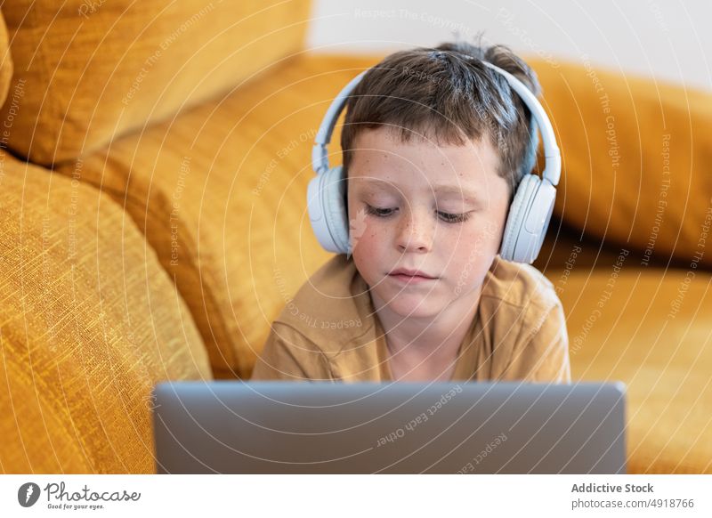 Boy lying on a sofa with headphones on his head and using the laptop child computer boy caucasian technology male kid serious concentrate cartoon movie indoors
