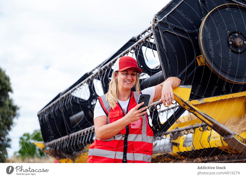 Smiling woman having phone call near combine harvester talk smartphone agriculture worker conversation countryside smile friendly content speak cheerful uniform