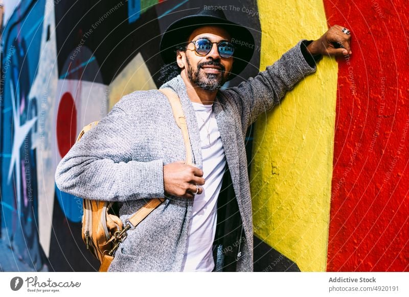 Stylish Hispanic man leaning on graffiti wall smile street style positive touch urban building male adult hispanic ethnic appearance happy backpack trendy