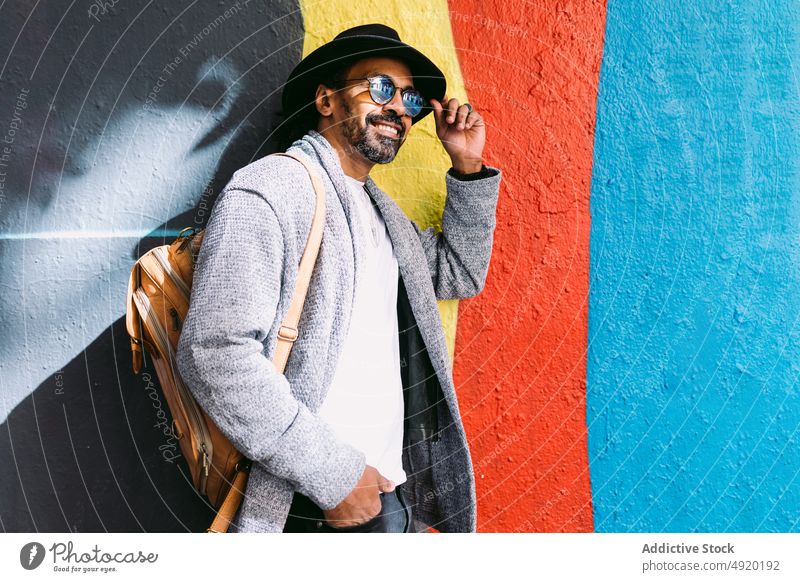 Stylish Hispanic man leaning on graffiti wall and touching his hat smile street style positive urban building male adult hispanic ethnic appearance happy