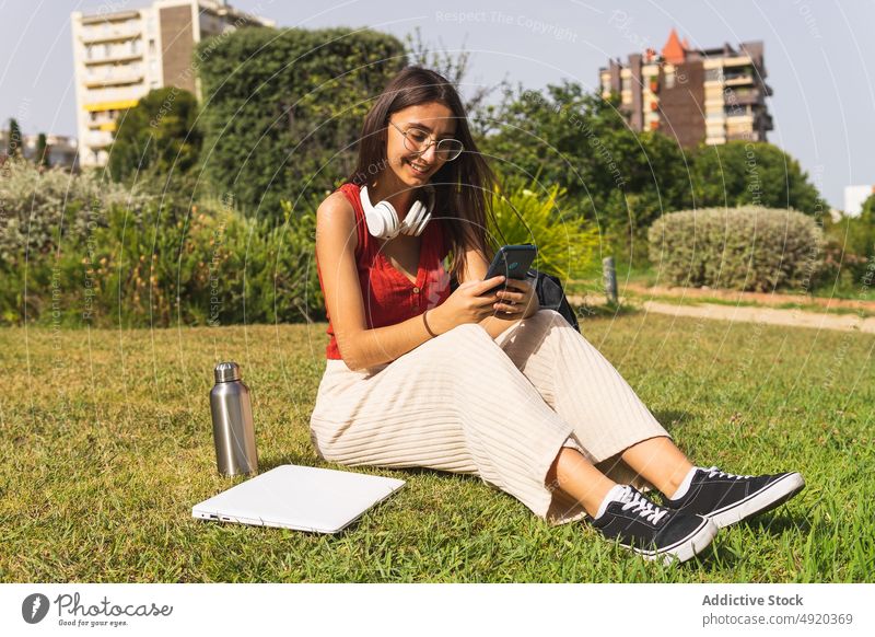 Smiling woman browsing smartphone on lawn rest text message park online social media chat app chill free time scroll positive joy smile cheerful student city