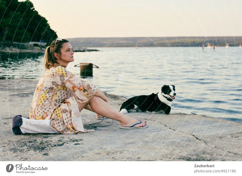 Woman in summer dress sitting with her dog by the sea Summer dress tourist Dog Ocean look Evening tranquillity To enjoy Meditative Think Friends Love of animals