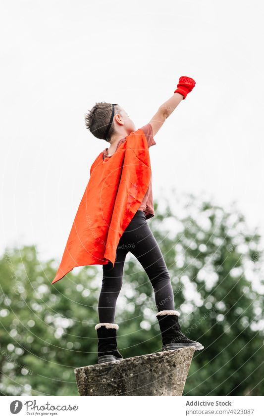 Anonymous superhero in hedgehog mask in park with arms raised with clenched fist boy play climb costume pretend kneel stone block courage child kid cape