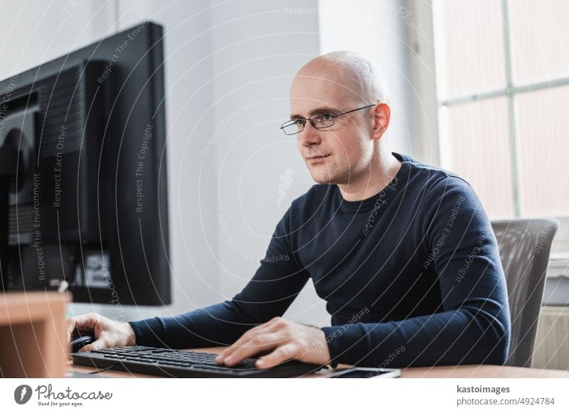 Handsome Young Businessman Wearing Eyeglasses Sitting at his Table Inside the Office, Looking at the Report on his Computer Screen businessman monitor portrait