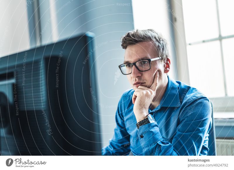 Handsome Young Businessman Wearing Eyeglasses Sitting at his Table Inside the Office, Looking at the Report on his Computer Screen. businessman monitor portrait