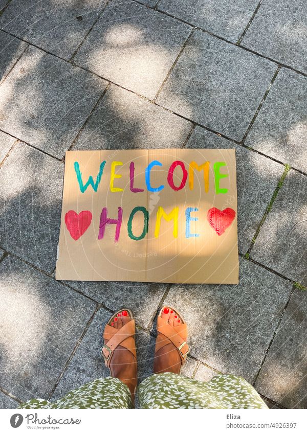 Woman stands in front of a sign on which is written in colorful letters "Welcome Home welcome home variegated Return writing authored feet Travel Return