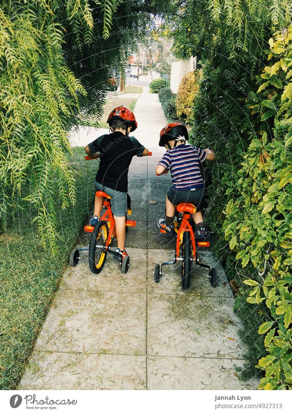 Two little brothers riding their bikes with training wheels and helmets on suburban neighborhood sidewalk Two boys riding bikes child riding bike