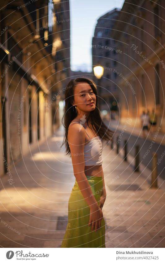 Beautiful young Asian woman walking the street of European historic town at night with streetlights on. Female tourist from China traveling in Europe. Fashion and style concept.