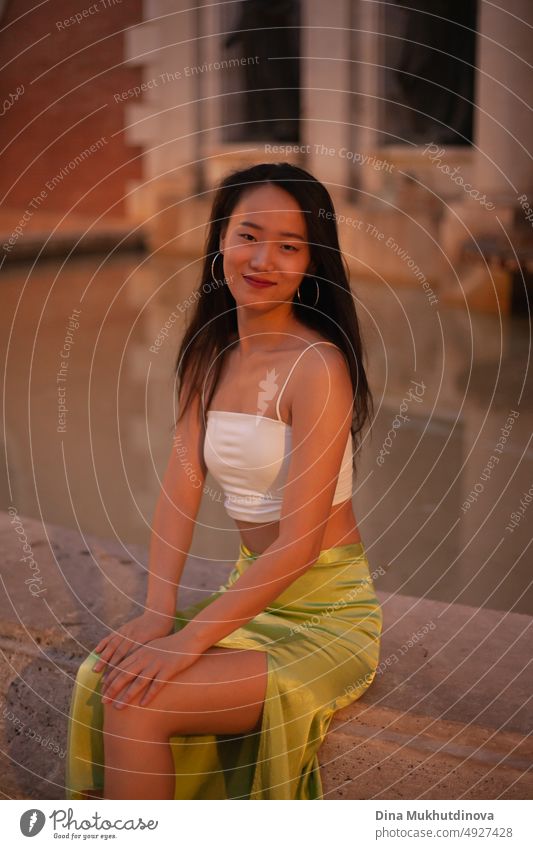 Beautiful young Asian woman sitting near fountain in European historic town at night with streetlights on. Female tourist from China traveling in Europe. Fashion and style concept.