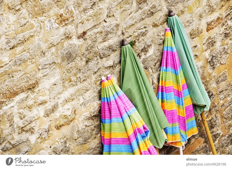 two green and three colored striped closed parasols leaning against an old stone wall / end of season Sunshade Closed Summer shade dispenser Stone wall
