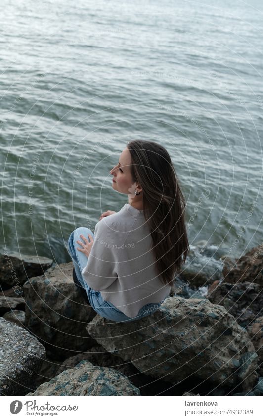 Young woman sitting on the rocks at the wild beach during outcast weather hair relaxing recreation young outdoors female sea shore coastline waterfront portrait